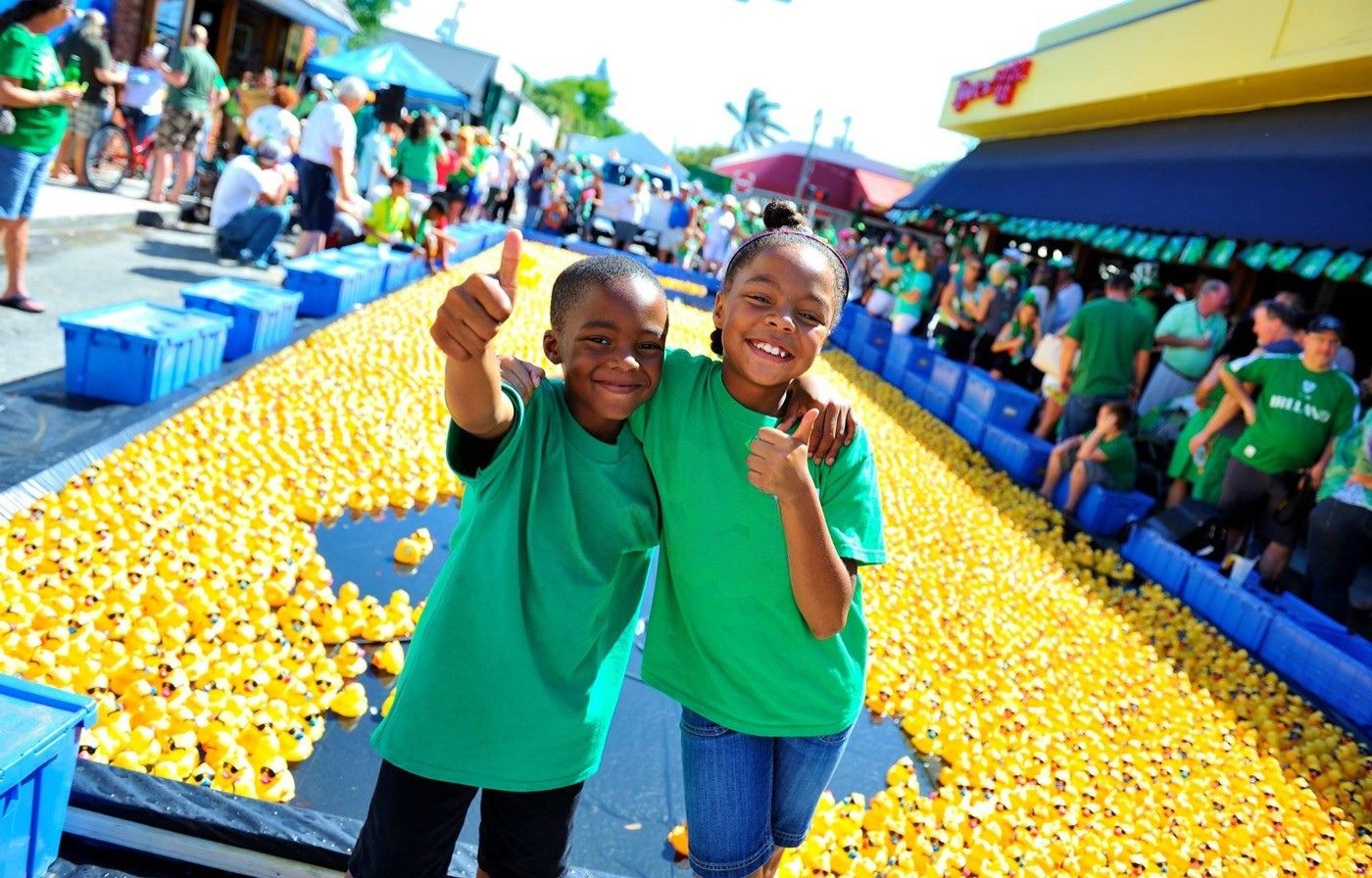 Two smiling kids in green shirts give a thumbs up beside a long lane of yellow rubber ducks at a community duck race.