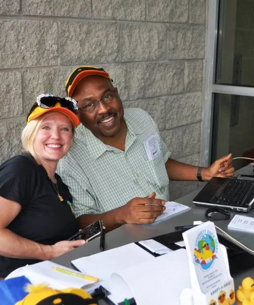 Two smiling volunteers staff a duck-race registration table.