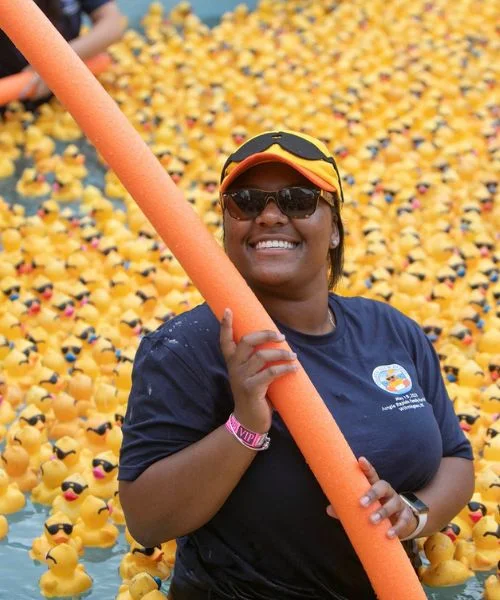 A volunteer guides rubber ducks down the water with a pool noodle.